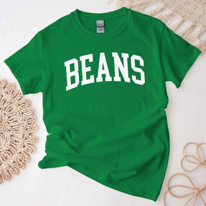 Puede incluir: Camiseta verde de manga corta con texto blanco que dice "BEANS" en una fuente universitaria.