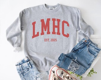May include: A heather gray sweatshirt with the letters "LMHC" in red, with "EST. 2025" below. The sweatshirt is paired with blue jeans and white sneakers with red accents.