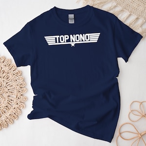 以下が含まれることがあります： 白文字で「TOP NONO」と書かれたネイビーブルーのTシャツ。星と水平線でデザインされています。Tシャツは白い表面に平らに置かれ、背景には装飾的な要素があります。