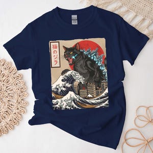 May include: A navy blue short-sleeve t-shirt featuring a graphic print of a black cat resembling a monster emerging from a large ocean wave near a city. The design includes a red sun, blue glowing spikes, and Japanese text in a vertical white box.