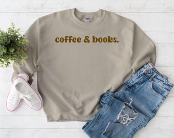 May include: A tan sweatshirt with the text "coffee & books" in brown lettering. The sweatshirt is paired with blue jeans and white sneakers with red trim. A small green plant is also in the image.