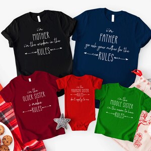 Op de afbeelding: Familie bijpassende t-shirts in zwart, marineblauw, bordeauxrood, rood en groen. Elk shirt heeft een witte tekst die luidt: "I'm Mother", "I'm Father", "I'm the Older Sister", "I'm the Younger Sister" en "I'm the Middle Sister".