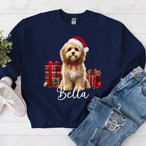 May include: Navy blue sweatshirt featuring a Cavalier King Charles Spaniel wearing a Santa hat and a red bow. The dog is surrounded by wrapped gifts and ornaments. The name "Bella" is written in white script below the dog.