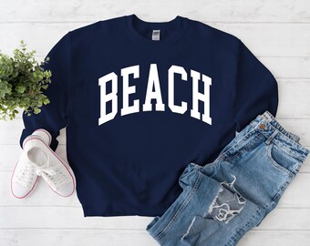 May include: Navy blue sweatshirt with the word "BEACH" in large, white, arched lettering. The sweatshirt is paired with light-wash denim jeans and white canvas sneakers with red accents. A small green plant is also in the image.