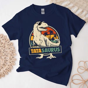 May include: A navy blue t-shirt with a retro-style graphic of a dinosaur wearing sunglasses and a palm tree background. The text "TATASAURUS" is printed below the dinosaur.
