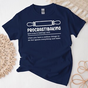 May include: A navy blue t-shirt with a white graphic design that says "PROCRASTIBAKING [pro-cras-ti-baking] verb when you have a million things to do but ignore everything and bake."