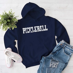Peut inclure: Un sweat à capuche bleu marine avec un motif blanc qui dit "Pickleball" avec une silhouette de personnes jouant au pickleball.