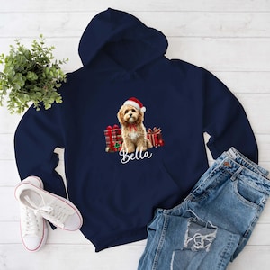 Puede incluir: Sudadera con capucha azul marino con un gráfico de un perro con gorro de Papá Noel y pajarita roja, con el nombre "Bella" en escritura blanca. El perro está rodeado de regalos envueltos. Zapatillas blancas y vaqueros azules también en la imagen.