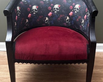Puede incluir: Sillón con marco negro con asiento de terciopelo burdeos y respaldo con estampado de calaveras y flores. El asiento está ribeteado con encaje negro. La silla tiene un acabado de madera oscura.