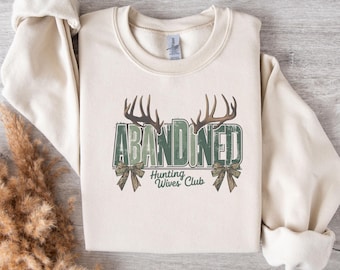 Puede incluir: Sudadera color crema con la palabra "ABANDONED" en letras verdes, coronada por astas marrones. Debajo, se lee "Hunting Wives Club". Dos lazos verdes flanquean la palabra. La sudadera tiene mangas largas.