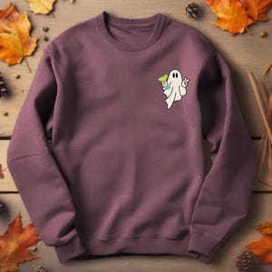 May include: A mauve-colored sweatshirt featuring a small embroidered ghost holding a green cocktail and giving a peace sign. The ghost is white with black eyes. The sweatshirt is made of a soft material.