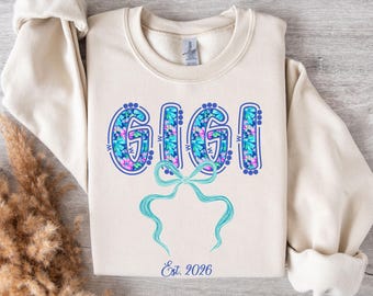 May include: Cream-colored sweatshirt with the word "GIGI" in a floral design with blue and pink accents. A light blue bow is below the word, and the text "Est. 2026" is at the bottom. The sweatshirt has long sleeves.