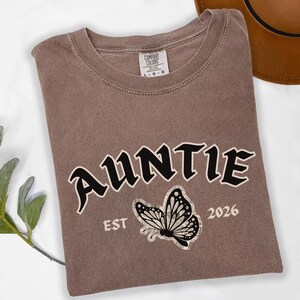 May include: A brown Comfort Colors t-shirt with the word "AUNTIE" in black, outlined in white, and a butterfly graphic. Below the butterfly is "EST 2026". The shirt is folded on a white surface.