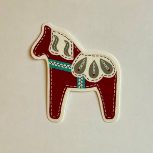 May include: A maroon Dala horse sticker with white, teal, and gray accents. The horse features a decorative design with a stitched outline and a white border. The sticker is a folk art design.