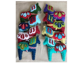 Peut inclure: Une collection de huit ornements en feutrine en forme de cheval de Dala, déclinés en vert, bleu, rouge, violet et jaune. Chaque cheval présente des motifs cousus décoratifs, des selles contrastées et des broderies blanches. L'image inclut le texte Pearl and Poppy, Scandinavian Felt Ornament Pattern et Dala Horse.