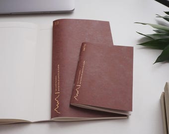 Puede incluir: Tres cuadernos marrones de diferentes tamaños están colocados sobre una superficie blanca, uno de ellos abierto para revelar páginas en blanco. Las cubiertas presentan texto dorado y una línea decorativa. Un portátil y vegetación son visibles en el fondo.