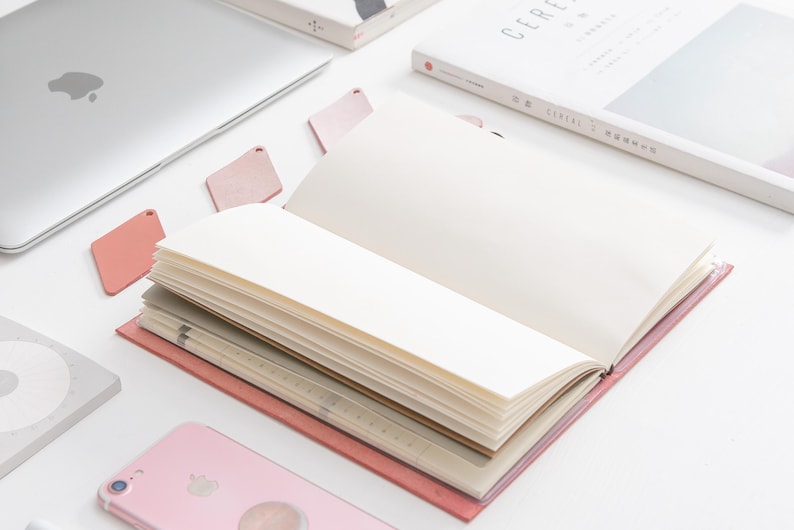K&ouml;nnte beinhalten: Ein offenes rosa Notizbuch mit leeren Seiten liegt auf einer wei&szlig;en Oberfl&auml;che. Das Notizbuch ist umgeben von anderen B&uuml;roartikeln, darunter ein Laptop, ein Telefon und ein Buch.