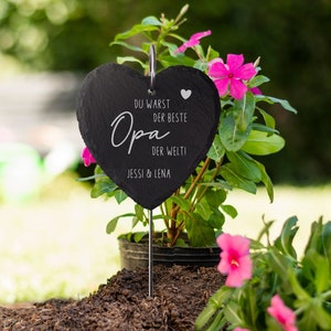 K&ouml;nnte beinhalten: Schwarzes herzf&ouml;rmiges Schiefer-Schild mit dem Text "Du warst der beste Opa der Welt" und "Jessi & Lena" in wei&szlig;. Das Schild ist an einem Metallstab befestigt und in einen Topf mit rosa Blumen gesteckt.