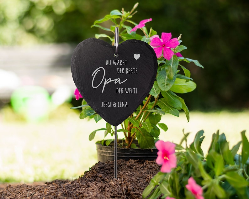 K&ouml;nnte beinhalten: Schwarzes herzf&ouml;rmiges Schiefer-Schild mit dem Text "Du warst der beste Opa der Welt" und "Jessi & Lena" in wei&szlig;. Das Schild ist an einem Metallstab befestigt und in einen Topf mit rosa Blumen gesteckt.