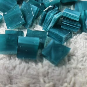 May include: A collection of small, square, teal-colored glass tiles scattered on a soft, white, textured surface. The tiles have a slightly translucent quality, with variations in color and texture.