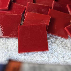 May include: A pile of small, square, red glass tiles. The tiles are smooth and shiny, and they have a slightly textured surface.