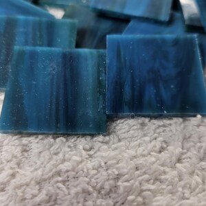 May include: A collection of square, blue glass tiles with a marbled appearance. The tiles are various shades of blue and are resting on a white, textured surface. These tiles are suitable for mosaic art projects.