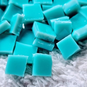 May include: A close-up shot of numerous square, turquoise-colored glass tiles. The tiles are scattered and slightly overlapping, creating a textured surface. The background is a soft, white, fuzzy material.
