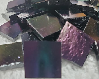 May include: A collection of iridescent, square-shaped glass tiles. The tiles have a dark, reflective surface that shifts in color, displaying hues of purple, green, and blue. The tiles are scattered, creating a textured, mosaic-like appearance.