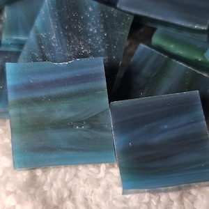 May include: A collection of square, glass mosaic tiles in shades of blue and teal. The tiles have a marbled appearance, with darker hues blending into lighter ones. The tiles are scattered on a white, textured surface.