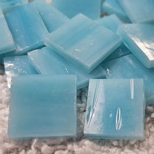May include: Close-up of light blue square glass tiles. The tiles are arranged in a scattered pattern, with varying angles and slight variations in color. The tiles have a smooth, glossy surface, and are likely for mosaic or craft projects.