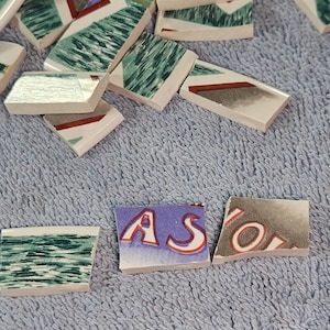 May include: Scattered ceramic tile fragments with various designs. Some tiles feature green, white, and red geometric patterns. Others display the letters "ASO" in a stylized font. The tiles are on a blue textured surface.