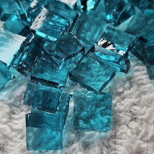 May include: Close-up of scattered, translucent, square-shaped glass tiles in shades of teal and aqua. The tiles are arranged on a soft, white, textured surface. The glass appears to have a slightly rippled texture.