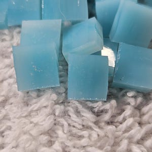 May include: Close-up of several light blue, square-shaped glass tiles. The tiles are scattered on a white, fluffy, textured surface. The tiles appear to be translucent and have a slightly irregular shape.