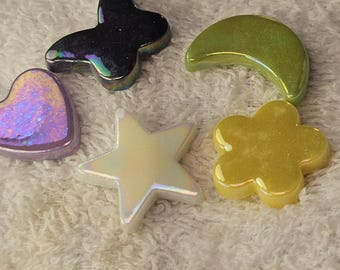 May include: A collection of small, smooth, decorative resin shapes in various forms including butterflies, crescent moons, hearts, stars, and flowers. The pieces feature diverse finishes such as iridescent, glittery, solid, and pearlescent colors.