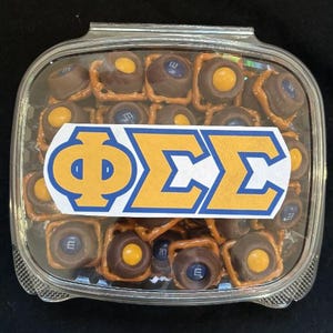 May include: A clear plastic container filled with chocolate-covered pretzels and peanut butter cups. The container has a yellow and blue sticker with the letters "ΦΣΣ" on it.