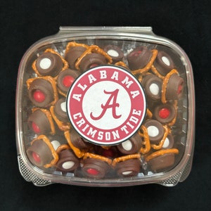 May include: A clear plastic container filled with chocolate-covered pretzels. The pretzels are topped with white and red candy. A red and white circular sticker with the text "Alabama Crimson Tide" and the letter "A" in the center is visible.