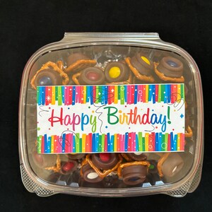 May include: A clear plastic container filled with chocolate-covered pretzels and colorful candies. A colorful birthday card with the text "Happy Birthday!" is attached to the container.