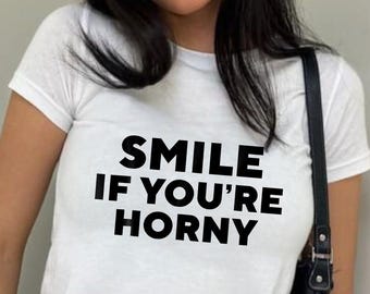 May include: White t-shirt with the text "SMILE IF YOU'RE HORNY" in black block letters. The shirt is paired with blue jeans and a black shoulder bag.