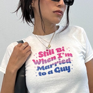May include: White t-shirt with the text "Still Bi When I'm Married to a Guy" in pink and blue. The shirt is short-sleeved and worn by a person wearing sunglasses and a necklace. A black purse is held in the left hand.