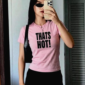 May include: A light pink short-sleeved t-shirt featuring the text "THATS HOT!" printed in bold black capital letters across the chest. The shirt is paired with a black shoulder bag and a silver-toned necklace with a cross pendant.