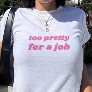 May include: White t-shirt with the phrase "too pretty for a job" in pink and orange lettering. The shirt has a crew neck and short sleeves. A black shoulder bag is visible on the left side of the image.