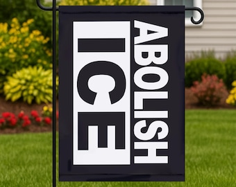 Op de afbeelding: Een zwarte tuinvaandel met de woorden "ICE" en "ABOLISH" in grote witte blokletters. De vlag hangt aan een zwarte metalen standaard in een grasrijke tuin, met een huis en struiken op de achtergrond.