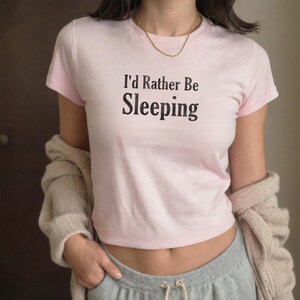 Pode incluir: Um top cropped rosa claro com a frase "I'd Rather Be Sleeping" em preto. A camisa tem mangas curtas e é combinada com calças de moletom cinza. Um cardigan bege está drapeado sobre os ombros.