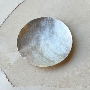 May include: A silver, round, shallow dish with a textured surface. The dish is made of metal and has a slightly hammered finish.