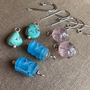 May include: A collection of jewelry components including two teardrop-shaped turquoise beads, two cylindrical blue textured beads, two round translucent pink beads with dark inclusions, and three silver-toned metal earring hooks with loops.