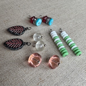 May include: A collection of handmade jewelry components, including leaf-shaped charms with pink accents, red and blue bead charms, clear glass beads, pink glass beads, and green and white beaded bars. The components are arranged on a neutral fabric surface.