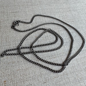 May include: A dark gunmetal gray curb chain necklace featuring interlocking oval links. The necklace includes a small circular spring ring clasp at one end, allowing for secure closure. The metal has a smooth, polished finish that reflects light.
