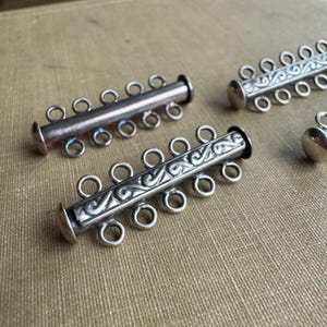 May include: Close-up of several silver-toned jewelry clasps. Each clasp has a cylindrical bar with multiple loops for attaching jewelry components. Some clasps feature an engraved design. The clasps are on a neutral-toned fabric background.
