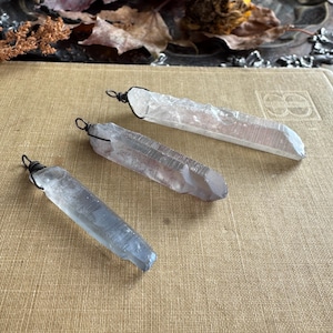 May include: Three raw crystal pendants with wire wrapping. The crystals are various shades of clear, gray, and blue. The pendants are displayed on a textured, tan surface, with dried leaves in the background. The crystals are approximately 3-4 inches long.