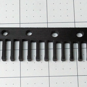 May include: A black comb with a series of teeth and several circular holes along the top edge. The comb is rectangular and appears to be made of a solid material. The teeth are evenly spaced and extend downward.